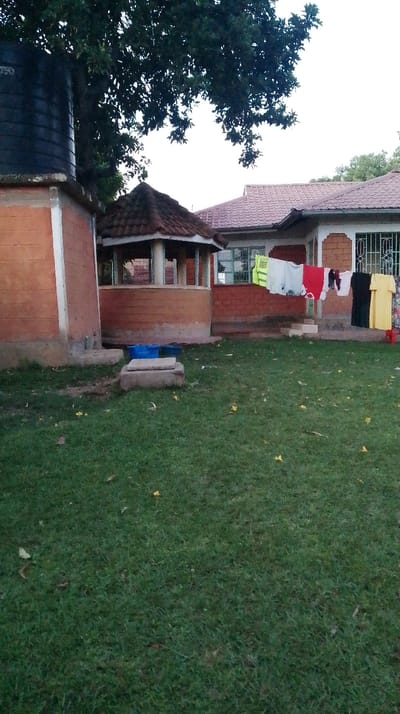 This image captures an outdoor residential area in Siaya Township ward, Kenya. The foreground consists of a well-maintained green lawn with scattered yellow foliage. A rectangular concrete slab is centrally located on the grass. To the left, a black cylindrical water tank rests on a reddish-brown brick base. Adjacent to it, a hexagonal gazebo with a reddish-brown tiled roof and open sides is visible. Two blue plastic containers are positioned on the ground near the gazebo's base. In the background, a single-story house with a reddish-brown tiled roof features white plastered walls accented with reddish-brown brick sections. Multiple windows with white security grilles are apparent. A clothesline extends across the front facade of the house, displaying various items of laundry, including a yellow safety vest, white shirts, a red shirt, a dark shirt, and a yellow dress. A small red stool supports the clothesline near the yellow dress. Dense green tree foliage occupies the upper left portion of the frame, set against a light, uniform sky. No individuals are present in the scene.