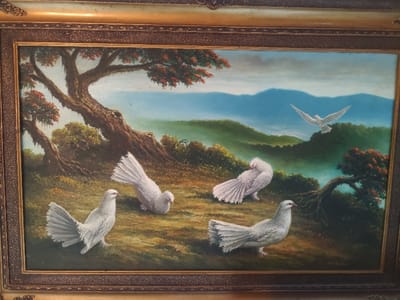 The image displays an oil painting set within an ornate, gold-colored frame. The depicted scene is a hilly, verdant natural landscape, representing a rural or semi-rural area in Kokui, Russia. In the foreground, four white doves are positioned on a ground covered in brown and green vegetation. One dove on the far left stands facing right with its tail fanned. To its right, another dove is hunched, facing left, appearing to peck at the ground with its tail fanned. Centrally, a third dove faces left while preening its feathers, its tail fanned outwards. A fourth dove, situated in the bottom right, stands facing left with a fanned tail.
A large tree with a thick, gnarled trunk and reddish-orange foliage dominates the left midground. Another tree with similar characteristics, its trunk bent low, is visible on the right midground. Rolling green hills recede into the background, culminating in distant blue mountains under a light blue and pale gray sky. A fifth white dove is depicted in flight in the upper midground, wings outstretched, heading towards the upper right of the frame. A dark, illegible signature is present in the lower right corner of the painting.