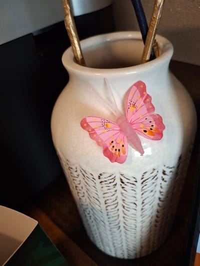 An ornate white ceramic vase with a crackle glaze finish sits on a dark wooden surface. The vase is decorated with an intricate raised pattern in a darker color. A pink fabric butterfly with black and orange spots and clear antennae is attached to the front of the vase. Inside the vase, two thin, metallic gold-colored objects and a dark blue object, possibly decorative sticks or brushes, are upright. In the background, a dark, indistinct object, possibly an appliance, is visible to the left. The scene is lit from the front and right, casting shadows. The image appears to be a close-up shot of the vase and its contents. There is no information or visual cue in the image that indicates the location is in Lēdmane, Latvia.
