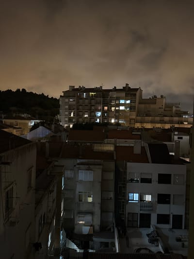 This is a nighttime, eye-level, medium shot of a dense urban residential area in Algés, Portugal. The scene is dominated by buildings of varying heights and architectural styles, illuminated by interior lights and ambient city glow against a dark, cloudy sky. In the background, a tree-covered hillside rises, silhouetted against the hazy sky.

The foreground and midground are filled with buildings. To the left, a dark building with visible window bars and a fire escape is partially obscured by shadow. Adjacent to it is another building with several illuminated windows, some with visible blinds or curtains. Further to the right, a complex of buildings with terracotta tiled roofs is visible. One building in this cluster has a lighter facade with multiple windows, some of which are lit.

In the mid-to-background, a tall, multi-story apartment building with a grid-like facade stands out. Its numerous windows are lit with a mixture of yellow and blueish light, suggesting occupancy. The building has balconies and visible structural elements. Behind and to the right of this building, other apartment blocks with varying window configurations and balconies are visible, receding into the distance.

The overall impression is one of a typical European city neighborhood at night, characterized by dense housing, illuminated windows, and a somewhat muted atmosphere due to the cloudy conditions. No people are visible in the frame.