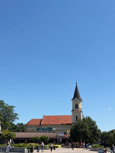 The image captures a street scene in Siófok, Hungary, under a clear blue sky. The focal point is the tall, yellow and white church steeple topped with a black spire and clock face. The church's red-tiled roof is visible, attached to a beige building.
People are walking and standing along the sidewalk in front of the building, partially obscured by trees. Signage for a restaurant or shop, "Basta Grill Restaurant Siófok," is apparent. The shot is taken from a low angle, looking up at the church and sky. The image is rotated 90 degrees clockwise.