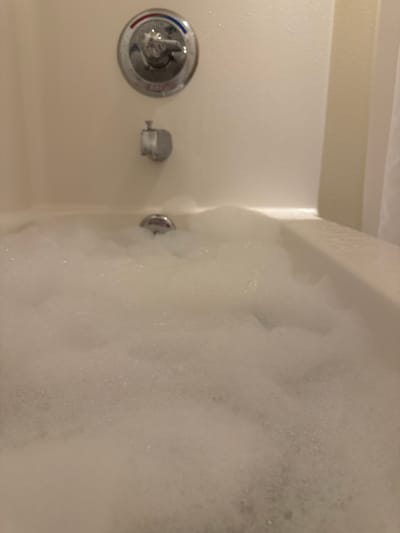 The image shows a close-up view of a bathtub filled with soap bubbles. The bathtub is white, and the bubbles obscure much of the scene. The bathtub fixtures, including the water temperature control and shower diverter, are visible on the left side of the frame. There is no evidence to determine the location in the city of Anchorage, United States of America. The scene includes a bathtub and soap bubbles.