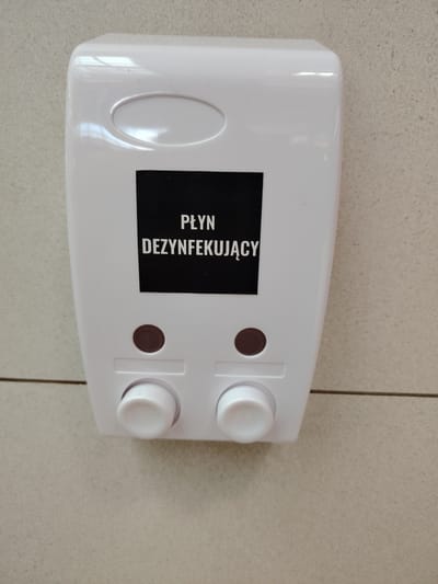 The image shows a white plastic automatic soap or sanitizer dispenser mounted on a wall with tiled beige and gray surface. The dispenser has a smooth, curved top with an oval indentation and a black label with white text in Polish that reads "PŁYN DEZYNFEKUJĄCY," which translates to "DISINFECTANT LIQUID." Below the label, there are two circular brown buttons or indicators, and beneath those, two white dispensing knobs. The overall setting appears to be a public restroom or a common area in a building where hand hygiene is emphasized. The lighting suggests it is daytime, and there are no specific weather cues. The image is a close-up of the dispenser, so no people or further environmental details are visible.