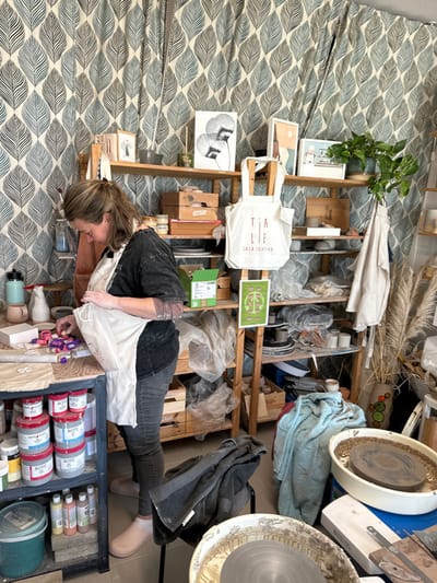 The image depicts a pottery studio or creative workshop located in Mafra, Portugal. A single adult female, with light brown hair pulled back, is observed leaning over a dark-framed workbench in the left midground. She wears a dark t-shirt, dark jeans, and a light-colored apron, which is smudged with light-colored material, likely clay. Her forearms also show traces of clay. She is actively manipulating small, colorful objects, including pink and purple pieces, on the workbench surface, alongside a white rectangular block.
The workbench's lower shelves contain multiple containers of glazes or paints, predominantly labeled "BOTZ," alongside smaller bottles of liquids.
Behind the individual, a multi-tiered wooden shelving unit extends across the background, filled with various pottery-related items. These include ceramic pieces (bowls, mugs, vases, unfired clay objects), wooden crates, and items wrapped in clear plastic. A white tote bag, printed with "TIALLE CASA CRIATIVA," hangs prominently from one of the shelves. Decorative elements on the shelves include framed artworks, such as a monochromatic floral print, abstract pieces, and a potted green plant on the top right. A poster with green circular graphics is affixed to a shelf support.
In the lower right foreground, two white plastic tubs, likely slop buckets or pottery wheel basins, are visible, one containing a dark pottery wheel bat. A dark grey towel is draped over a black stool, and light blue fabric is crumpled nearby. A tall, patterned container on the far right holds dried pampas grass-like decor. The wall behind the shelving unit is covered with wallpaper or fabric featuring a repeating pattern of stylized blue-green and dark grey leaves on a light background. The space is brightly lit.