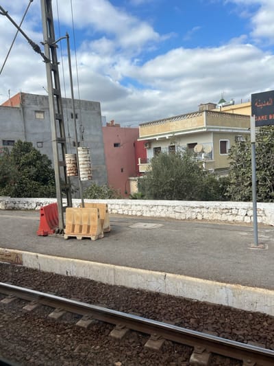This image depicts a static scene at the train station (Gare de Sidi Slimane) in the city of Sidi Slimane, Morocco, under a partly cloudy sky.

In the immediate foreground, a single set of railway tracks with dark gravel ballast and grey concrete sleepers runs horizontally across the bottom of the frame. Above the tracks, a light grey concrete platform edge, exhibiting minor cracking, parallels the railway.

The midground features an asphalt-surfaced platform. Centrally positioned is a tall, dark grey metal utility pole, labeled "293-016", supporting overhead lines and two sets of heavily corroded, cylindrical counterweights. Adjacent to this pole are three portable barriers: two beige, interlocked, concrete or heavy plastic blocks with a ridged design, and one smaller red plastic barrier. Further to the right on the platform, a slender grey metal post holds a black rectangular sign displaying white text; partially visible are the Arabic word "المدينة" (The City), "SIDI SLIMANE", and "GARE DE". A low, white-painted stone retaining wall extends across the mid-distance, separating the platform from vegetated ground.

The background comprises a cluster of buildings and dense foliage. From left to right, above the vegetation, a multi-story, unpainted grey concrete building with small, rectangular windows is visible. To its right stands a two-story, reddish-pink building. Furthest right is a two-story yellowish building featuring a decorative, patterned parapet, a small balcony with a wrought-iron railing, several windows, and two white satellite dishes affixed to its exterior. A small green finial or dome-like structure protrudes from the roofline of the yellowish building. Green trees and thick bushes partially obscure the lower portions of these background structures. No individuals are present, and no actions are discernible.
