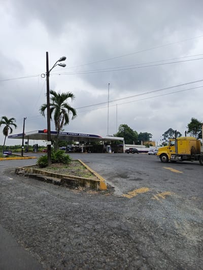 The image depicts a gas station in Quevedo, Ecuador under an overcast sky. The gas station, labeled "Nibal Fonfay Barzola," is visible in the background, with multiple gas pumps under its canopy. Various vehicles, including cars and a large yellow semi-truck, are present in the fore and middle ground. A concrete island with a light pole, small palm trees, and other vegetation is in the foreground. Yellow painted arrows are visible on the paved surface, directing traffic flow. A tall communication tower is situated behind the gas station, adding to the scene's industrial character. The surrounding environment shows signs of vegetation and structures typical of a roadside location.