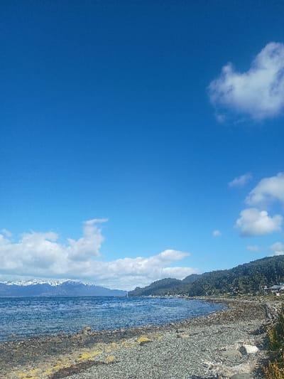 A vibrant blue sky dominates, punctuated by scattered white clouds. A shoreline stretches along the right side of the frame, featuring a gravel beach and a body of water. Lush green vegetation covers the land adjacent to the beach. Mountains rise in the background, partly obscured by clouds.