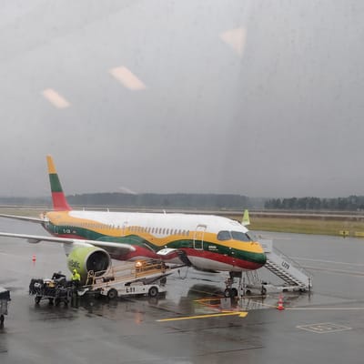 An airBaltic Airbus A220-300 aircraft, registration YL-CSK, is parked on a wet tarmac under an overcast sky at what is identified as Skulte, Latvia. The aircraft features a distinctive green, yellow, and red livery, consistent with the Lithuanian flag. Ground handling operations are underway on the left side of the aircraft. A person wearing a high-visibility yellow jacket is standing near a black baggage cart loaded with various bags and packages, positioned adjacent to the left engine. A white motorized baggage loader, labeled "L11", connects the baggage cart to the open front cargo hold of the aircraft. A set of mobile passenger stairs, marked "L229", is positioned at the aircraft's front left door. An orange and white safety cone is placed on the tarmac near the nose landing gear. The tarmac displays yellow directional arrows and parking position markings. In the background, a tree line is visible beyond the runway/taxiway, appearing hazy under the gray sky. The image exhibits reflections of interior lighting fixtures (two rectangular and one triangular) and minor surface imperfections, indicating it was captured through a window.