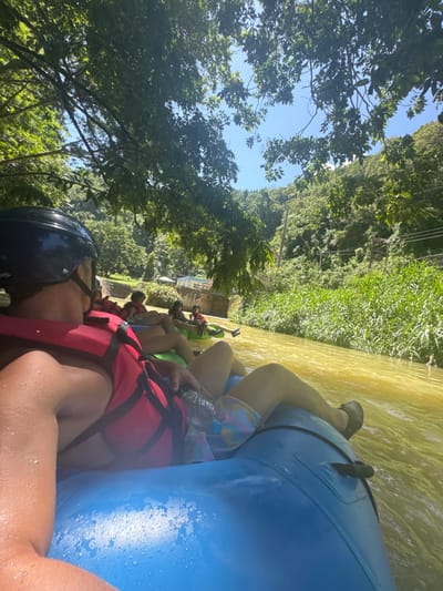 A group of individuals is engaged in a river tubing activity in Woods Town, Jamaica. The foreground shows a person from a first-person perspective, wearing a black helmet, a red life vest, and patterned shorts, seated in a blue inflatable tube with visible water droplets. A clear plastic water bottle is held near their lap. Behind them, several other participants are visible in similar inflatable tubes, some green, also wearing life vests, floating down a murky, yellowish-brown river. The riverbanks are densely lined with tall green vegetation, including what appears to be reeds or sugarcane on the right side. In the background, a multi-arched stone or concrete structure, possibly a bridge or aqueduct, spans the river on the left, with small buildings nestled among abundant tropical foliage and distant green hills. Overhead, large tree branches with lush green leaves partially obscure the bright blue sky, which contains no clouds. Utility poles and wires are visible among the trees on the right bank.