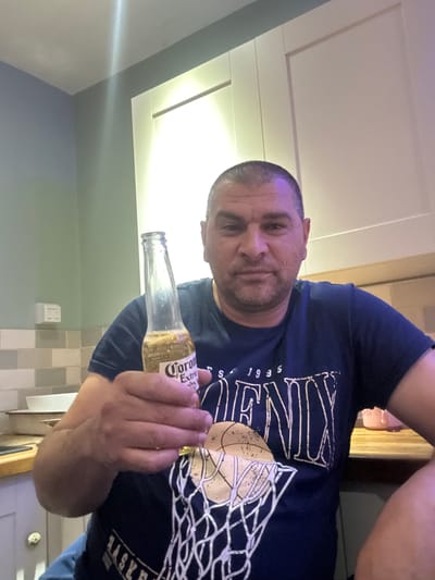 A man is shown holding a bottle of Corona Extra beer while sitting in what appears to be a kitchen. He is wearing a dark blue t-shirt with a graphic design that includes the word "PHOENIX" and a basketball net. The background shows kitchen cabinets and tiled walls. The lighting suggests it is indoors, possibly evening. There is no indication this scene is in Boston, United Kingdom.