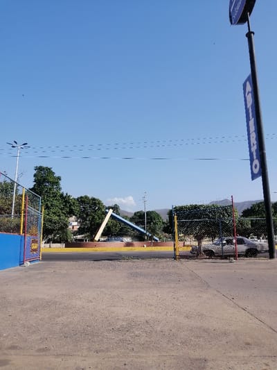 A sunlit scene reveals a concrete lot leading to a fenced area with colorful posts. Beyond the fence stands a modern blue and yellow sculpture set against green trees and distant hills under a clear sky. A silver car is parked near the fence on the right. A tall black pole with a blue "ABIERTO" sign dominates the right side of the image. Overhead power lines stretch across the sky.