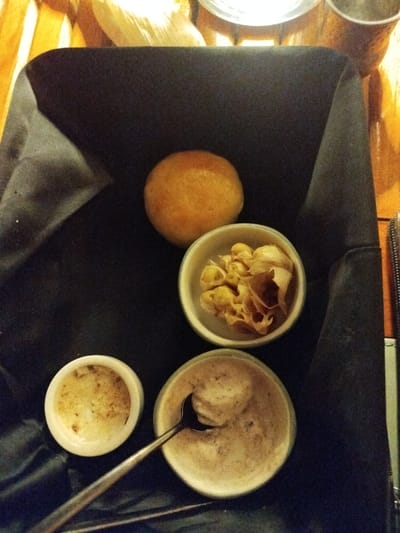 A close-up, high-angle shot shows a dark woven bread basket containing a round bread roll, a small white bowl filled with light-colored, brittle-looking food items, and two smaller white bowls, one with a spoon resting in it. The bread roll is positioned in the upper center of the basket. The bowl with the food items is to the right of the bread roll. The two smaller white bowls are in the lower portion of the basket, with the one on the right containing a spoon. The background appears to be a wooden table. The scene is located in Kendhoo, Maldives.