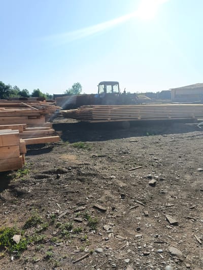 The image depicts an outdoor lumberyard on a sunny day. In the foreground, a large pile of cut lumber is visible, stacked horizontally. Behind this pile, a tractor is parked next to another large stack of lumber, which appears to be on a flatbed trailer supported by round logs. To the right, a wooden structure, possibly a building or shed, is partially visible. The ground is covered in dirt, gravel, and scattered debris. Trees and foliage form a backdrop beyond the lumberyard. The overall impression is of an active work or storage area for wood products, likely located in Strelka, Russia, given the prompt's context. No people are directly visible in the image.