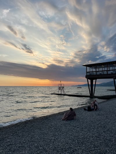 An eye-level, vertical, outdoor shot captures a twilight scene at the Krayevsko-Armyanskoye beach in Russia. The sky is a canvas of dramatic clouds, transitioning from pale blues and whites at the top to hues of orange and yellow near the horizon, indicating the sun is setting. A vast, choppy body of water stretches across the middle of the frame, with gentle waves lapping onto a densely pebbled beach. In the foreground, three individuals are seated on the beach. On the left, a person in a patterned dress sits facing away from the camera. Further to the right and slightly behind, a couple sits close together, with one person leaning back. A backpack rests on the ground between them.

Dominating the right side of the midground is a modern, dark-colored structure on stilts, resembling a restaurant or observation deck. It features a glass-enclosed upper level illuminated by strings of small lights. A narrow pier extends from the beach towards the structure. On the left side of the midground, a metal structure with two life preservers and a yellow flag stands on a short pier extending into the water. In the distance, a small boat is visible on the water, and a hazy shoreline with distant buildings and mountains forms the backdrop. The overall atmosphere is serene, with the natural beauty of the sky and sea juxtaposed against the man-made structures.
