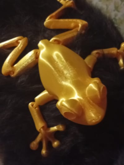 A close-up, high-angle shot shows a golden, articulated toy frog lying on a dark, furry surface. The frog's body is plump and rounded, with visible seams from its 3D printed construction. Its limbs are extended, and its webbed feet are splayed. The lighting creates bright highlights on the frog's smooth surface. The background is predominantly dark and textured, suggesting a soft material like fur or plush. There is no visible indication of the city of Berezivka, Ukraine, or any people or specific actions occurring.