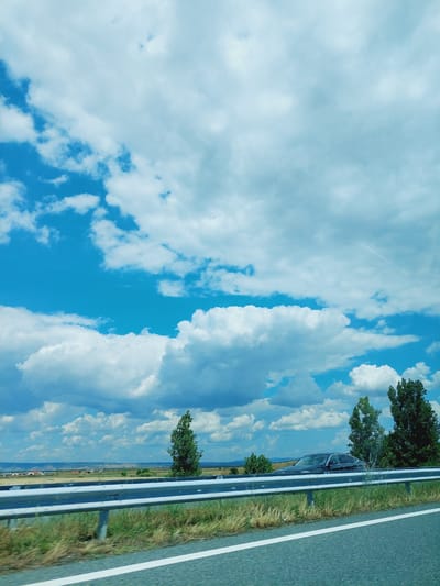 The image depicts a scenic view from the side of a road, likely in Zheleznik, Bulgaria, characterized by a vast expanse of blue sky dotted with fluffy white clouds. A metallic guardrail runs horizontally across the foreground, separating the road from a grassy verge. Beyond the verge, a flat landscape unfolds with sparse vegetation and a few scattered trees. In the middle ground, a dark-colored car is visible, partially obscured by foliage. The overall impression is one of a serene, possibly rural, roadside environment under a bright, partly cloudy sky.