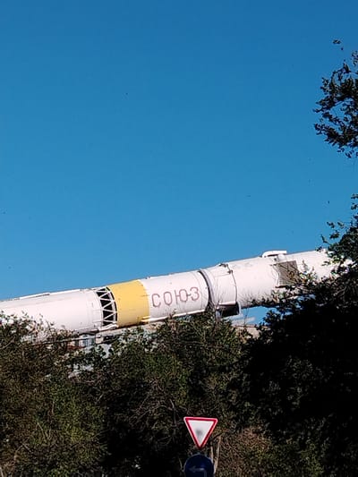This image, taken from a low angle looking upwards, features a section of a white rocket with a yellow band and some red Cyrillic lettering. The rocket is positioned horizontally, with parts of it obscured by lush green trees in the foreground. The sky above is a clear, bright blue, suggesting a sunny day.
The red text on the rocket reads "СОЮЗ-3", which translates to "Soyuz-3". This indicates the object is likely a representation or remnant of a Soyuz spacecraft or launch vehicle, a significant part of Soviet and Russian space exploration history.
At the bottom of the frame, a yield sign is visible on a pole, with a directional arrow sign below it. These elements suggest an outdoor setting, possibly near a public area or exhibition related to space exploration. The presence of trees and the clear sky indicate it's daytime, likely morning or afternoon. There are no people visible in the image. The overall mood is one of quiet observation, focusing on the imposing technological artifact against a natural backdrop. The location context of Baikonur, Kazakhstan, a historic and active space launch site, strongly reinforces the interpretation of this object as a space-related exhibit or monument.