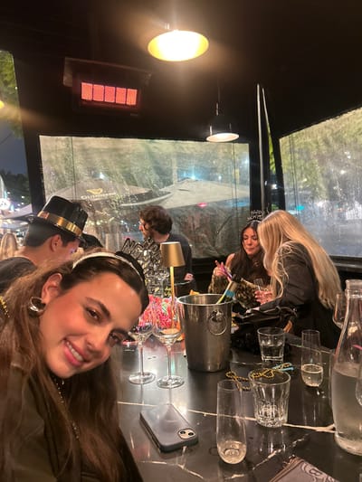 A group celebrates, possibly New Year's Eve, in an outdoor dining area enclosed by transparent barriers. A woman in the foreground smiles for a selfie, wearing a jeweled headband. Champagne is being poured. Another woman with long blonde hair is also pouring it. A man in a top hat is partially visible. A heat lamp is on the side of the dining area. Glasses, a champagne bucket, and a smartphone are on a dark marbled table. The background shows an outdoor setting at night.