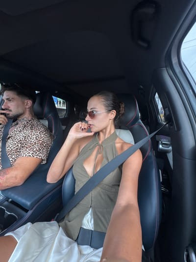 Two people are inside the back of a dark-colored vehicle. One person, a woman, is seated wearing a green top, sunglasses, and a seatbelt. The other person, a man, is lying down wearing a leopard-print shirt and seatbelt. The location appears to be Khok Kloi, Thailand based on a building visible through the car window. The vehicle has a sunroof.