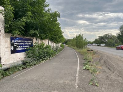 A paved pathway curves to the right alongside a concrete wall and dense green trees. A banner on the wall reads in Kazakh, "We repair bumpers of any complexity." Beyond the trees, a two-lane road is visible with several cars driving. The sky is overcast with gray clouds.
