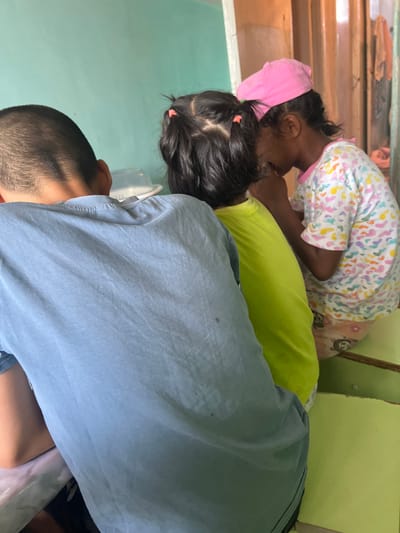 Three young children are seated together in an indoor environment, likely a dwelling. The child in the foreground, viewed from the back, wears a light blue t-shirt. To their right, another child in a bright lime green t-shirt leans forward. The third child, facing towards the right and partially obscured, wears a white t-shirt with a colorful heart pattern and a pink cap. The background consists of a light turquoise wall and wooden vertical paneling. The scene appears to be a candid moment captured in Şamaldı-Say, Kyrgyzstan, suggesting a domestic or community setting.