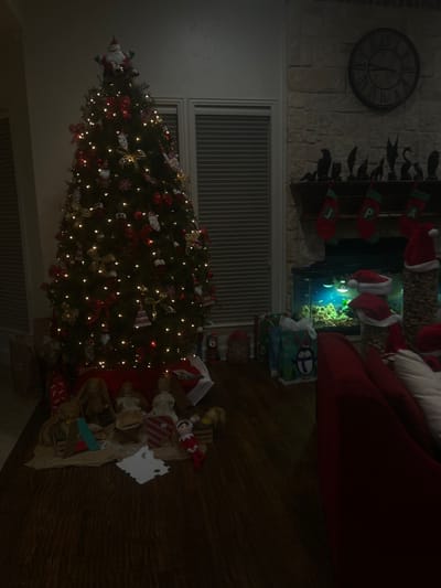 A decorated Christmas tree stands brightly with ornaments and lights near a fireplace adorned with stockings. Presents are stacked beneath the tree and around the base, with a large red couch nearby. A clock hangs on the wall above the fireplace, near a wrought iron decoration. The room is dimly lit, creating a cozy holiday ambiance.