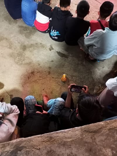 This image, taken in Kecamatan Cimaung, Indonesia, presents an overhead view of a group of children and young people gathered in what appears to be an indoor or semi-outdoor setting with a concrete floor. The dim, artificial lighting suggests the scene takes place in the evening or at night.

In the foreground, several individuals are seated on the ground. One child, wearing blue jeans and a dark top, is actively using a smartphone held horizontally, seemingly watching or playing a game. Beside another child's leg, a second smartphone with a red case is visible on the floor. A small, orange plastic object, possibly a toy, lies on the concrete floor in the mid-ground.

Further into the background, a line of several other children or young adults are seated, mostly seen from behind. Their attire varies, including a blue denim shirt, a black hoodie with partially legible white or light blue text on the back (appearing to be "ung-tee" or similar), a red, white, and black striped shirt, and a light blue or grey long-sleeved shirt. Some individuals are barefoot, while others wear footwear. The concrete floor shows signs of wear and staining.

The activity appears to be a casual gathering where the children are collectively engaged, with several focused on smartphones. A low wall or step in the immediate foreground indicates different levels within the space. The overall atmosphere is one of relaxed communal interaction and entertainment.