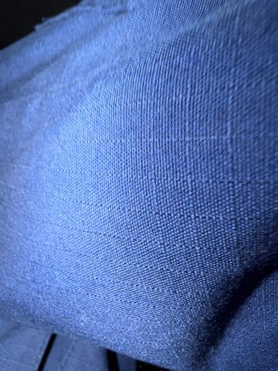 The image displays a close-up, abstract view of dark blue textured fabric, possibly denim or a similar material. The texture shows a distinct woven pattern with raised threads creating a ribbed effect. The lighting creates highlights and shadows across the fabric, emphasizing its three-dimensional qualities. The bottom of the image reveals a glimpse of black material with a linear pattern, suggesting it might be a seam or another part of the garment. There is no discernible urban environment or any indicators that this image is located in Shingak-Kul', Russia. The scene is entirely focused on the fabric's detail.