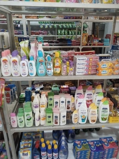 The image displays the interior of a retail establishment, likely a minimarket or small store, located in Aceh Harbor, Indonesia. The scene is dominated by multiple metal shelving units holding various consumer products.
From top to bottom, the visible shelves are organized as follows:
* The second shelf from the top features a prominent display of baby care products, including lotions, shampoos, and oils from brands such as Cussons Baby and Baby Happy, packaged in white, pink, blue, and yellow bottles. To the right, stacked boxes of Pepsodent toothpaste (specifically strawberry flavor) and Mizone Junior products are arranged.
* The third shelf showcases a wide assortment of adult body care products, including lotions and shampoos, predominantly in white, light green, pink, and yellow bottles. Brands such as Dalan D'Olive and Marina are identifiable, with some Marina products featuring fruit illustrations like strawberry, orange, and avocado. Behind these, smaller packaged items or soap bars are visible.
* The bottom shelf is dedicated to household cleaning supplies. Multiple blue and white bottles of Vixal toilet cleaner are displayed, alongside other liquid cleaning agents and sprays, including products like Wipol and Pristine. To the right, stacked boxes, possibly containing mosquito coils, are present, with "Bayam" visible on some packaging.
The products are neatly arranged on the shelves for sale. No individuals are present in the image. The environment is brightly lit, suggesting artificial lighting.