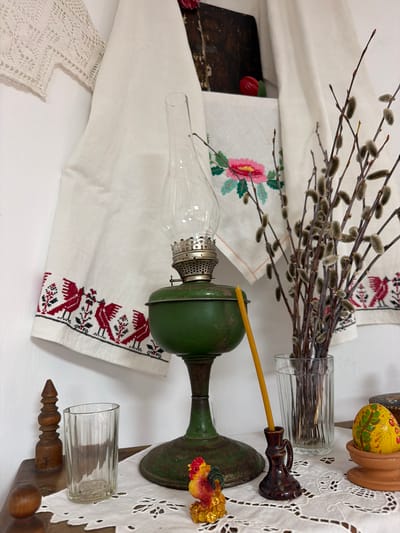 An indoor still life depicts traditional folk art and objects. In the background, a white textile with red embroidered birds and floral motifs hangs on a white wall, partially obscured by a dark, book-like object with a red item on top. To the right, a bouquet of pussy willow branches in a clear glass vase stands beside a white embroidered cloth. In the foreground, a green oil lamp with a glass chimney sits on a wooden surface covered with a white embroidered tablecloth. A yellow candle is placed in a small, dark brown ceramic holder next to the lamp. A small, decorative rooster figurine is positioned to the left of the candle. A clear glass with ribbed sides is to the left of the rooster, and a carved wooden finial stands behind it. A colorful, decorated egg sits in a terracotta holder on the right side of the frame. The scene is set indoors, likely in a domestic environment in Ust'-Kabyrza, Russia, evoking a sense of rural tradition and possibly related to Easter celebrations.