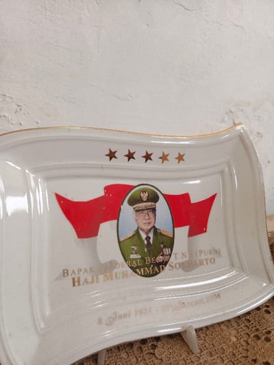 A commemorative porcelain plate features a portrait of Haji Muhammad Suharto, a general in the Indonesian National Army (TNI). Red and white imagery representing the Indonesian flag surrounds the portrait. Above the image, five gold stars are displayed. Below the portrait and flag, gold lettering reads "Bapak Jenderal Besar T.N (PURN) Haji Muhammad Suharto" and "8 Juni 1921 - 27 Januari 2008," likely referencing his birth and death dates. The plate has a gold rim and a wavy edge.