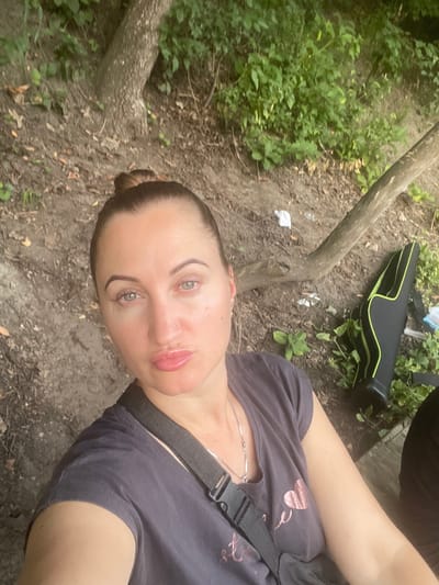 The image shows a woman with her hair in a bun, wearing a dark t-shirt with a heart design. She is taking a selfie outdoors, with a natural background of trees and foliage. A black and green case, possibly for a musical instrument or equipment, is visible to her right. The ground is covered in dirt, leaves, and some litter. The overall impression is of a casual, outdoor setting.