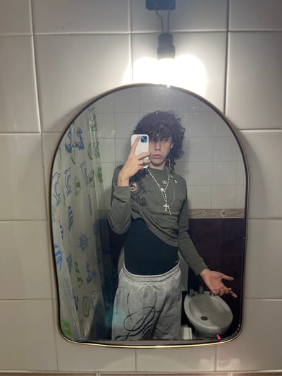 A mirror reflects a person with curly hair, wearing a green long-sleeve shirt, black undershirt, and gray sweatpants. The person is holding a white phone, presumably taking the picture. The reflection shows a bathroom setting with white tiled walls, a bidet, and a shower curtain with marine-themed drawings. The presence of a bidet suggests this location may be in Europe or South America. Based on the tiled walls and bidet placement, this is likely a bathroom in a residential building. Although specific city identification of Bahía Blanca, Argentina, is not possible solely from the image.