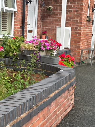 The image shows a close-up of a brick wall and garden outside a row of terraced houses in Wrexham, United Kingdom. The wall is constructed from red bricks, with a contrasting darker brick border along the top edge. Several flower planters are mounted on the wall, overflowing with a variety of colorful flowers, including pink, red, and white varieties. Lush green foliage surrounds the flowers, creating a vibrant and well-maintained garden display.
The setting appears to be an exterior residential area, likely a front garden or pathway leading to the houses. The brickwork and architectural style suggest a typical British terraced housing estate. The lighting indicates it is daytime, and the presence of wet foliage hints at recent rain or dew. No people are visible in the frame, focusing the attention on the garden and architectural elements. The overall impression is one of a neat and inviting home exterior. There is no visible text in the image.
