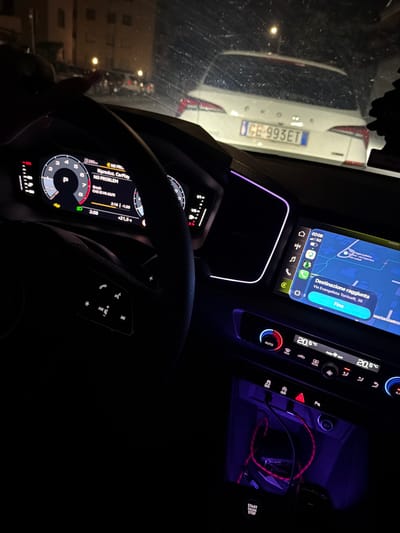 The image shows the interior of a car, specifically the dashboard and steering wheel, at night. The dashboard is illuminated with purple ambient lighting. The car's navigation system displays a route with the destination reached at "Via Evangelista Torricelli, 39" in Lissone, Italy. The driver's side dashboard shows the car is in park. A white car with the license plate "GE-993ET" is visible through the front windshield. The car's internal temperature is set to 20.5 degrees Celsius. The car's entertainment system is playing music from an artist named "Bresh" and a song titled "CHE 10 MI AIUTI". The image is taken from the driver's perspective.