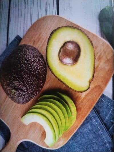The media file displays a close-up shot of avocados on a wooden cutting board. The subjects are two whole avocados, one halved and showing its pit, and several thin slices of avocado arranged in a fan shape. The avocados are placed on a light-colored, weathered wooden surface, possibly a table or countertop, with a blue cloth underneath the cutting board. The lighting suggests it might be daytime, with a bright but diffused light. There are no people visible, and the overall scene evokes a sense of freshness and preparation for a meal. The setting appears to be a kitchen or dining area, given the presence of the cutting board and cloth. No text is visible in the image.