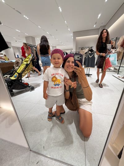 In the city of Alboraia, Spain, a woman and a young boy are taking a selfie in a brightly lit clothing store. The woman, on the right, is kneeling and holding up her phone to capture the reflection in a large mirror. She has dark hair and is wearing a green tank top and a white skirt, with a woven brown bag on her shoulder. The boy, on the left, is standing and smiling at the camera. He has dark hair and is wearing a white t-shirt with a graphic of a lion on a skateboard and grey shorts. Both are wearing headbands. In the background, other shoppers can be seen browsing the clothing racks. A yellow and black stroller is parked near them. The store's white tiled floor reflects the scene.