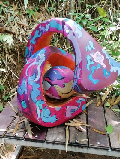 A colorful, abstract sculpture sits on a wooden table outdoors, surrounded by foliage. The sculpture is made of a pinkish-red material, decorated with swirling patterns in shades of blue, purple, and teal. It is a three-dimensional, curved form that creates an opening in the center, within which sits a spherical object. This sphere is also painted with similar swirling patterns in purple, blue, yellow, and pink. The wooden table appears weathered and is partially covered with fallen leaves. In the background, the scene transitions to a natural environment with lush green plants and trees, typical of the highlands of Da Lat, Vietnam. Sunlight filters through the leaves, casting dappled shadows on the sculpture and the surrounding ground.