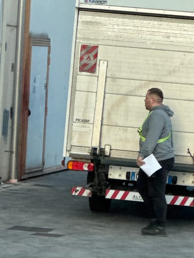 The scene depicts an outdoor industrial or commercial area in Sarcedo, Italy. A male individual stands in profile, facing right, next to the rear of a commercial truck. He is an adult, wearing a grey hooded sweatshirt with a yellow-green reflective safety harness, dark trousers, and dark shoes. His hair is cut short with a distinct shaved line visible on the side of his head. He holds a stack of white papers in his left hand.
The truck, positioned behind and to the left of the man, has a light-colored, ribbed rear panel. Markings on the truck include a partially visible "FURGOKIT" logo at the top, a red and white striped hazard sign featuring a white triangular 'A' symbol, and the word "PICASSO" printed on the lower left of the panel. The truck's rear features red taillights, a black structural bumper, and a prominent red and white striped reflective safety bar across the bottom, with the partial word "ALT" visible on a lower section. To the far left, a light blue building wall with a partially visible, dark-framed door is present. The ground is paved with asphalt or concrete, showing some rectangular dark patches. A vertical metallic pole is also visible on the far left.