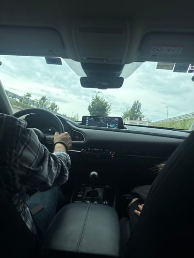 The image shows the interior of a car, looking forward from the passenger seat. A person's hands are on the steering wheel, and they are wearing a plaid shirt and blue jeans. The car's dashboard and infotainment screen are visible, displaying a navigation map. Another person, likely a passenger, is partially visible in the right side of the frame, wearing black jeans with a rip and a black jacket. The car is on a road with trees and a white fence visible outside the windows. The sky is cloudy. There are also several stickers on the car's windshield, including an airbag warning. The location is identified as Unknown, Mexico.