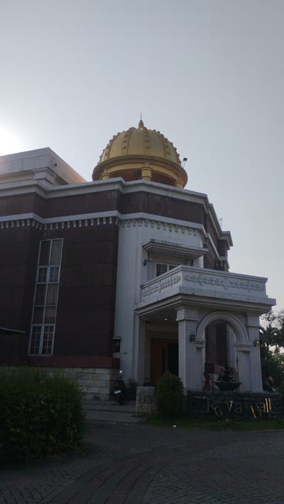 The image shows the exterior of a building with a prominent golden dome. The building has a dark brown facade with white trim and several windows. There is a white balcony with decorative carvings above an arched entrance. A sign on the ground in front of the building reads "Royal Hall". A motorcycle is parked to the left of the entrance. The area is paved with bricks. The sky is clear and bright. The building is likely located in Sidodadi, Indonesia, as indicated by the context.