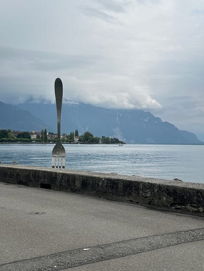An outdoor image depicts the monumental "The Fork" sculpture (La Fourchette) rising from the waters of Lake Geneva (Lac Léman) in Vevey, Switzerland. The polished metal fork, with four tines, stands prominently in the midground, partially obscured by a weathered, grey concrete retaining wall that runs horizontally across the lower portion of the frame. In the immediate foreground, a grey asphalt paved area, possibly a promenade or road, extends, showing a circular manhole cover and horizontal lines.

Beyond the sculpture, the calm, grey-blue expanse of Lake Geneva stretches to the horizon. Across the lake, a distant shoreline is visible, featuring low-lying buildings, green trees, and a small, dark boat afloat in the water. The background is dominated by large, dark blue-grey mountains, partially shrouded by heavy, low-hanging grey clouds that extend across the sky, indicating an overcast day. No people are visible.