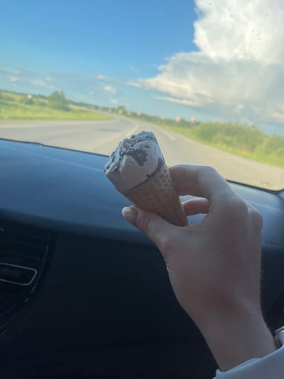 A person's hand holds an ice cream cone in the passenger seat of a car. The ice cream is light brown with dark brown chocolate chips. The car's dashboard is visible in the foreground. The background shows a road stretching into the distance with green trees and a blue sky with white clouds. There is no specific indication of the location being in Brodokalmak, Russia.