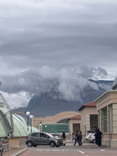 The image depicts a street scene in Milnerton, South Africa, with a view toward a mountain obscured by clouds. On the right side of the image is a multistory building with tiled roofing and architectural elements. Several people are walking on the sidewalk in front of the building. A gray car is parked on the street, and other cars are parked further down. In the foreground, there is a metal structure with lamps and possibly construction equipment. The sky is overcast.