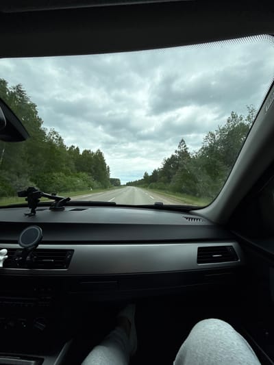 The image presents a view from inside a car, looking forward through the windshield onto a road. The perspective is tilted approximately 90 degrees clockwise, suggesting the photo was taken vertically.
The scene outside the car features a straight, two-lane asphalt road extending into the distance. Both sides of the road are densely lined with lush green trees and forest, indicating a rural or natural setting. The sky above is overcast with light grey clouds, suggesting a cloudy daytime, but with ample ambient light.
Inside the car, the dashboard and parts of the center console are visible on the right side of the frame. These include air vents with silver trim, and control panels with buttons. Two phone mounts are attached to the dashboard; one is a larger, multi-articulated mount, and the other is a circular magnetic mount clipped to an air vent. On the far right, a passenger's leg, dressed in light grey fabric, is visible, suggesting a passenger is seated in the front.
Visible text includes "AIRBAG" faintly discernible on the left A-pillar trim. On the car's control panel, words such as "FM", "AM", "MODE", and "SET" are visible, along with numbers "1" through "6" on buttons.
The image captures the activity of driving or being a passenger in a car during the daytime. The environment is consistent with a drive through a forested region, such as Beržiniai, Lithuania.