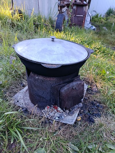 The image depicts an outdoor scene with a large, lidded cast-iron cauldron placed on a rustic, metal stove. The stove, which appears to be made from a repurposed drum, has an opening at the front containing glowing embers and ash, indicating it was recently used for cooking. The cauldron is silver and has a prominent knob on its lid. The entire setup is situated on a patchy, gray surface surrounded by overgrown green grass and some dried vegetation. In the background, out-of-focus elements suggest an industrial or abandoned area, with a weathered structure, a tire, and some metal debris visible. The lighting suggests it is either early morning or late afternoon, with sunlight casting long shadows and illuminating the grass. The image does not contain any people or indicate any specific activity beyond the presence of the cooking apparatus. The location is the city of Apraksin, Russia.