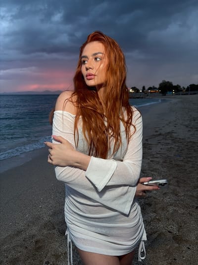 A woman with long red hair is on a beach at dusk in Alimos, Greece. She is wearing a white dress and holding a smartphone in her right hand, possibly taking a photo. The beach is sandy and the sea is visible in the background, with waves breaking on the shore. The sky is a mix of dark clouds and pink hues from the setting sun. The image is oriented vertically.