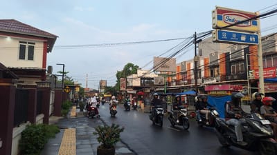 This is a street scene in Karawang, Indonesia, likely on a cloudy day, with wet asphalt indicating recent rain. A street is lined with various buildings, including a two-story white building with a red roof on the left, and multi-story buildings with orange and grey facades on the right. A prominent Indomaret convenience store sign is visible on the right. The street is busy with a flow of motorcycles carrying individuals, many wearing helmets. Some motorcycles are parked along the street. Buildings on the right appear to house shops and businesses, with some signs visible in Indonesian. There are utility poles with numerous wires crisscrossing the sky. A sidewalk with yellow tactile paving is present on the left side of the street. A potted plant with red flowers is located on the sidewalk. The overall atmosphere is that of an active urban commercial area.
