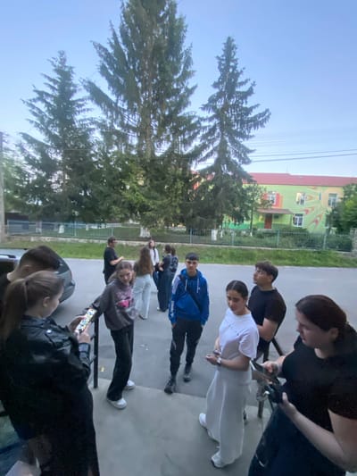 A group of approximately ten young people are gathered outdoors in what appears to be a residential area. They are standing on a sidewalk or paved area near a road. In the background, there are tall evergreen trees and a two-story building with a colorful, abstract mural on its exterior. The building's facade is predominantly light green with red and yellow accents. The sky is a clear blue.

The individuals are dressed in casual, modern clothing. Several are looking at their phones, while others are engaged in conversation. One person in the foreground is wearing a black leather jacket and holding a smartphone. Another is wearing a blue hooded sweatshirt. A person in a white t-shirt and long white skirt stands out among the group.

The overall scene suggests a casual social gathering, possibly before or after school, or during a break. The environment is typical of a suburban or small-town setting in Ukraine. No explicit indicators pinpoint this specific location in Tovstoluh, Ukraine, beyond it being a general depiction of such a place.