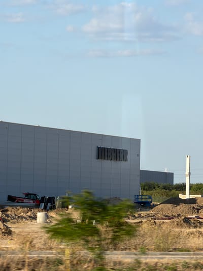 The image depicts a construction site in the city of Benkovski, Bulgaria. A large, modern warehouse-style building with "LIEBHERR" signage is prominent in the center. The building has a light gray, panelized exterior. In the foreground, construction activity is visible. A red Manitou telehandler is parked on a pile of dirt. Various construction materials, including concrete pipes and possibly scaffolding, are scattered around the site. A blue scissor lift is also present. The ground is uneven, with mounds of soil and dry vegetation. In the background, more construction appears to be underway, with another dark-colored building visible. The sky is a clear blue with a few scattered clouds. The overall impression is one of ongoing industrial development.